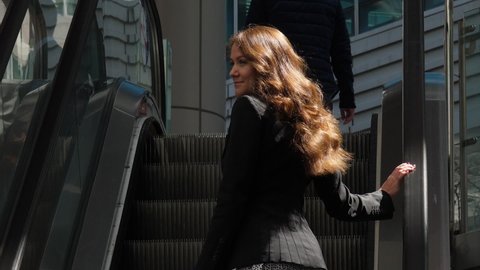 Woman move up, standing at outdoor escalator, look back, slow motion portrait shot. Sun light fall on long curly hair, shaded urban area at modern business district of Seoul
