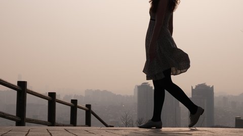 Woman walk by, legs against city in haze, staircase flight at Namsan mountain park. Dusty air hang over metropolis. Tourist lady wear light summer dress