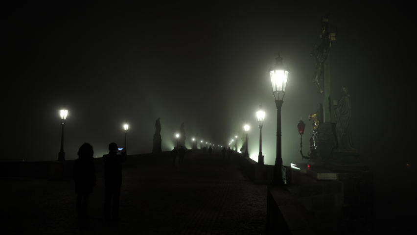 static shot close to lanterns and static view of statues and street lights on Charles Bridge and fog at night and silhouettes of pedestrians on cobblestone pavement on bridge and wind blowing in stor Royalty-Free Stock Footage #1067950721