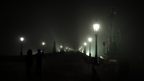 static shot close to lanterns and static view of statues and street lights on Charles Bridge and fog at night and silhouettes of pedestrians on cobblestone pavement on bridge and wind blowing in stor