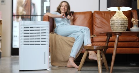 Woman switching on portable air conditioner while sitting on a couch at home. Enjoying fresh and clean air at home. Home appliances for cleaning and air conditioning