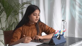 Motivated concentrated young Indian student studying on online courses.Young Asian girl university student using tablet computer distance learn lesson with teacher remote teaching on internet at home.