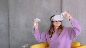 Young caucasian woman sitting on the armchair and using VR headset, holding controllers and looking in virtual reality.