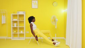 Young stylish woman is sitting on yellow chair in front of fan, trying to chill out in hot yellow room, feeling stuffy and tired, enjoying fresh air stream, Slow motion.
