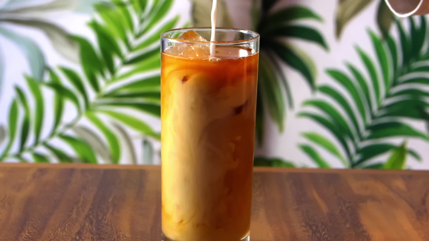 Smooth close-up of hand pouring cream into cocktail on wooden table with tropical leaves wallpaper as background. Glass of creamy alcoholic beverage on wooden surface. Drinks preparation