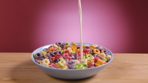 Milk is poured into a white bowl of cornflakes in form of multicolored rings in slow motion. Crispy colored breakfast cereal rings on a pink background. Dry healthy breakfast close up.