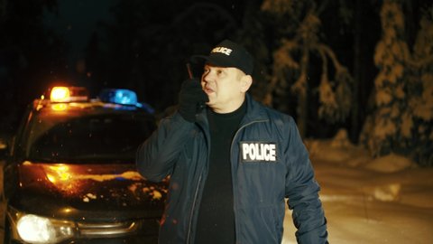 The policeman reports the details of the night's incident to the police station via a walkie-talkie. A police SUV with flashing lights stands behind on a country road in a snowy forest