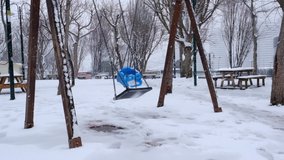 Empty swings are swinging in slow motion in a snowy park and snow is falling