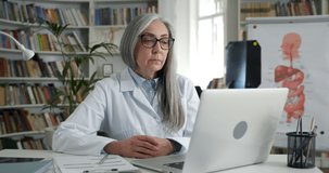 Lady in glasses and white professional rob talking and gesturing while looking at screen. Female mature doctor having online consultation while sitting at table in medical office.