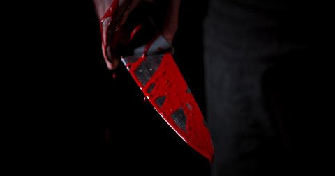Red blood flowing along the male hand with a sharp knife. Drops of blood fall down and luminating isolated on black background. Crime and violence concept. Blurred focusing revenge pattern