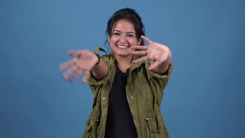 Young Asian friendly woman waving hand - hello. Greeting, say Hi to camera. Beautiful young trendy girl on blue studio background. portrait of happy adult girl  greeting showing symbol Hi.