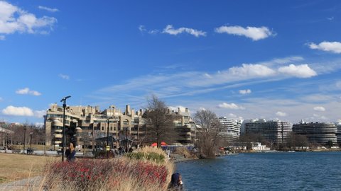 The Georgetown waterfront and Potomac River in Washington, DC on a winter day. The  Watergate Hotel and Kennedy  Center are seen in the distance. Blue skies and clouds overhead. The camera pans right.