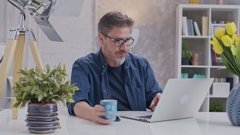 Bearded man working online with laptop computer at home sitting at desk. Home office, browsing internet, drinking coffee. Portrait of mature age, middle age, mid adult man in 50s.