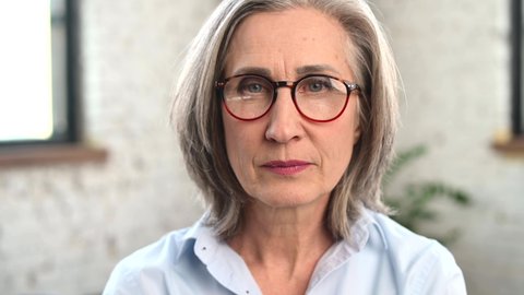 Close-up portrait of strict old senior business woman wearing smart casual shirt and stylish eyeglasses looks serious and then slimes friendly. A mature bossy lady, elderly purposeful leader