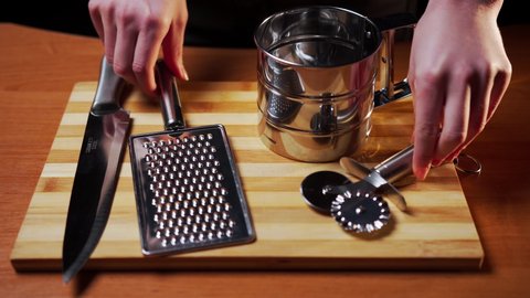 Female hands stack on a wooden board on the table metal kitchen utensils - a knife, a grater in the form of a platen, a sieve mug for flour and a double-sided knife for cutting pizza and dough