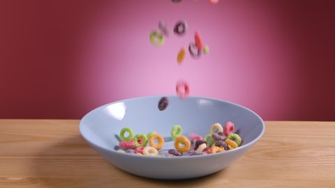 Breakfast cereal in form of multicolored corn rings fall and fill white bowl on table. Food close up on pink background. Colored cornflakes and dairy products. Rings are cereal. Slow motion.