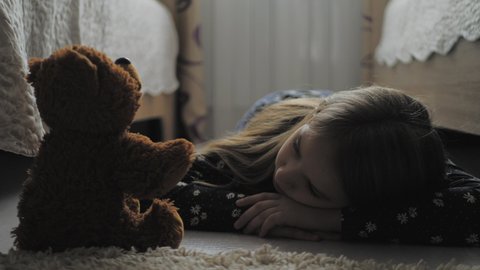 Depressed little girl and her best friend teddy bear in the dark gloomy room of his house..