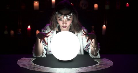 Young asian woman fortuneteller performing ritual with shining crystal ball. Gypsy girl cast spell and moving hands around ball psychic for prediction. Astrology, occultism and paranormal concept.