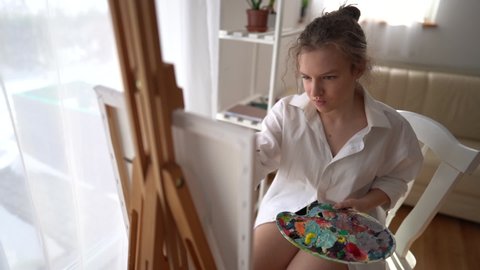 Schoolgirl learns to paint with oil paints. A girl sits in front of an easel and canvas and holds a brush and a palette with paints in her hands