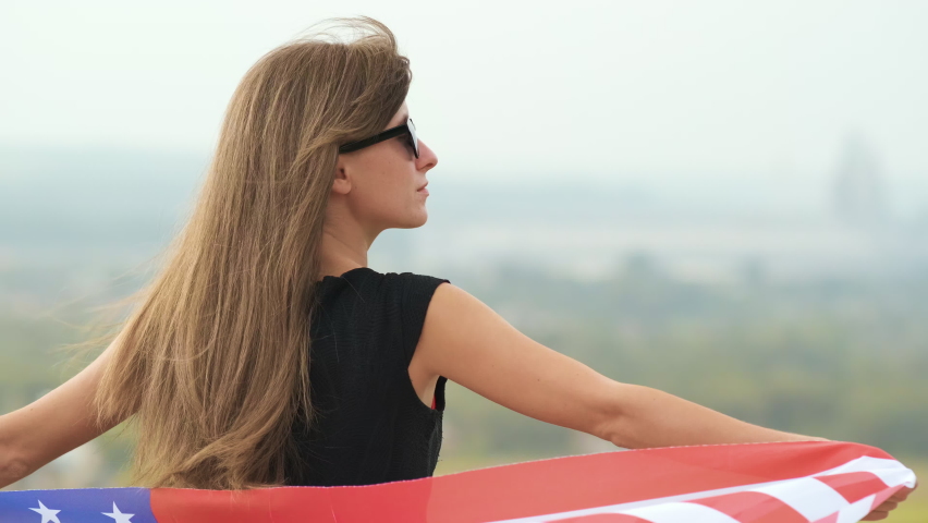 Young happy american woman with long hair holding waving on wind USA national flag on her sholders relaxing outdoors enjoying warm summer day.