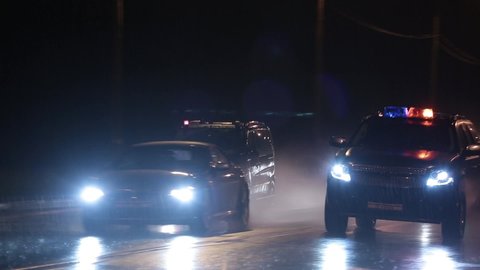 Police chase the intruder at night, cars drive by the camera at high speed through puddles. It's foggy and raining hard outside. Flashes of flashing lights and headlights.