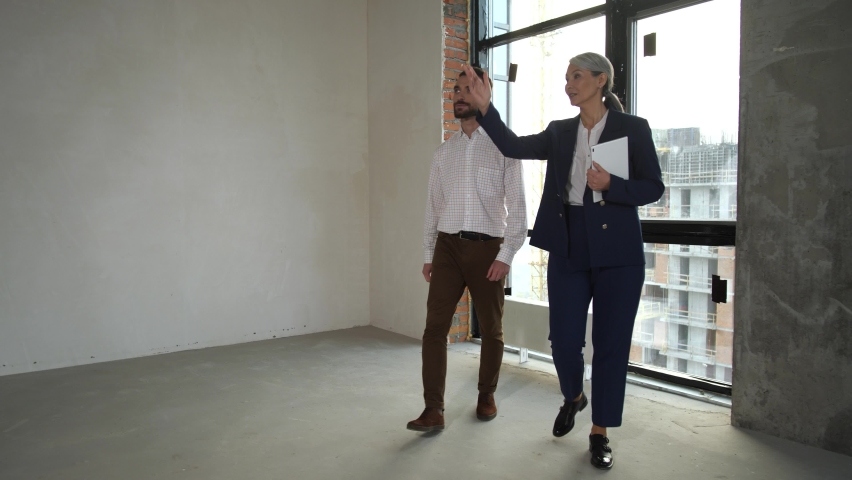 Delighted client listening to female real estate agent while walking around large empty space of apartment for sale. Professional realtor telling about future interior of property to buyer | Shutterstock HD Video #1068010007