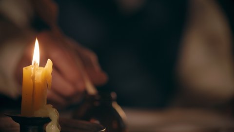 An 18th century scene focused on a lone candle in the foreground with a man using a quill pen out of focus in the background.