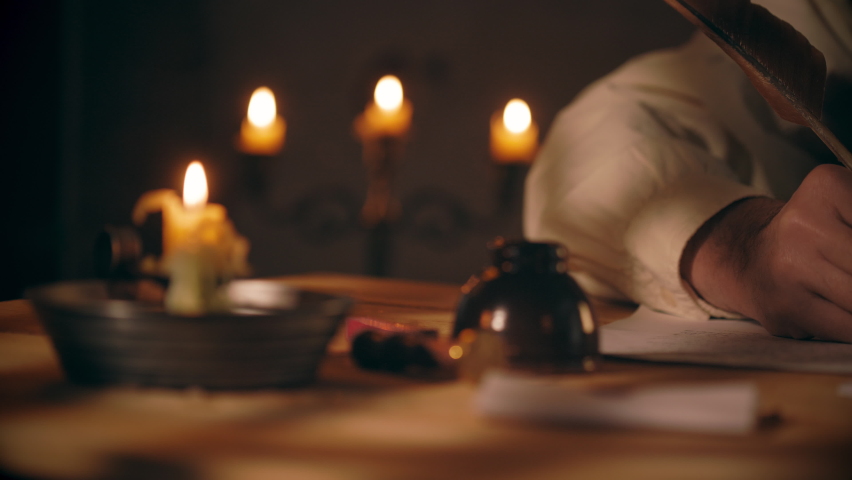 An 18th century scene tracks across a desk lit by candlelight and overhead window light of a man writing a letter using a feather quill pen. | Shutterstock HD Video #1068011003