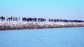 People in masks walk along rocky pier near azure ocean