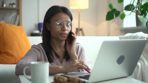 African woman with glasses at home working at computer and talking on phone Spbd. Black female model holds smartphone while typing on keyboard. There is a ring on finger, next to there is cup of