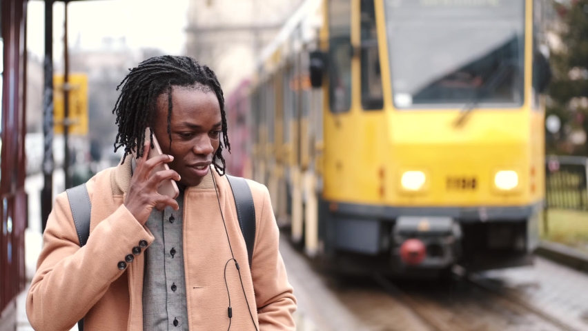 Young handsome african-american man with backpack talking with on cell phone while waiting at bus station.