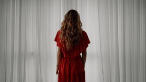 4K video of a young European girl in a red dress with long hair, standing in the house behind the window and opening window curtains with her hands in different directions