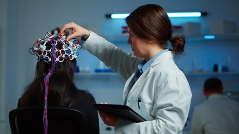 Woman patient wearing performant eeg headset scanning brain electrical activity in neurological research laboratory while medical researcher adjusting it, examining nervous system typing on tablet.