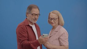 Old couple smiling and using cell phone over blue background. The couple showing each other the funny videos they watched.Technology addiction concept. 