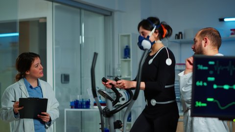 Doctor witing on clipboard health informations while patient with mask running on cross trainer and man medical researcher monitoring vo2, physical enducance, heart rate and psychological resistance