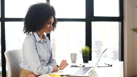 Mixed-race businesswoman in casual clothes with Afro hairstyle sitting in the office with panoramic window, studying or working online, having a virtual meeting, saying hello, waving at laptop screen