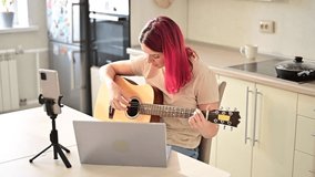 A woman sits in the kitchen during a remote acoustic guitar lesson. A girl learns to play the guitar and watches educational videos on a laptop