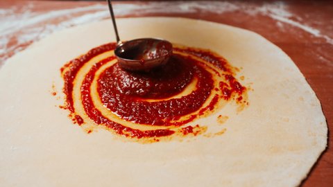 Chef is spreading Tomato Sauce on Pizza Dough with a Metal Ladle in Traditional Italian Pizza's Restaurant. Cooking traditional Sauce for Italian Pizza. Italian Food. Pizza's Making. Close up