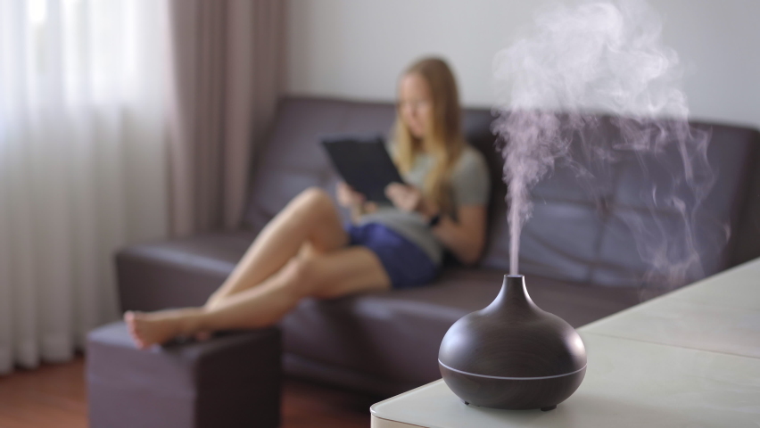 The woman in the background is sitting on a couch and a working aromatherapy diffuser is on a table in a foreground | Shutterstock HD Video #1068041633