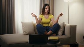 Calm young woman practicing yoga meditation poses at home. Beautiful lady doing online relaxation class, sitting on sofa in front of laptop.