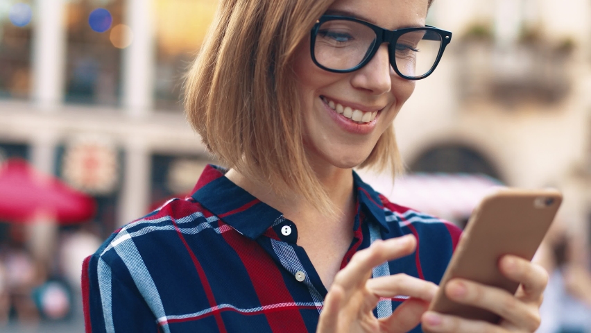 Close up of young blonde woman with glasses standing in the middle of street and chatting with friends using smartphone.