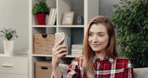 Young woman sitting at home on couch and talking to friends on video call holding smartphone and waving hand.