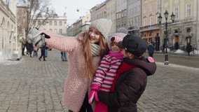 Lesbian couple tourists posing, taking selfie with adoption child girl kid on city street. Two women making photo or video chat with family and friends using smartphone during winter holidays vacation