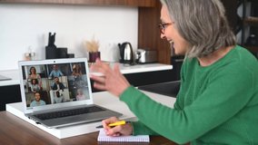 A senior mature gray-haired lady sits at the kitchen desk at home studying online on a laptop, watching a webinar with diverse group, participating in the online conference, taking a notes. Video call