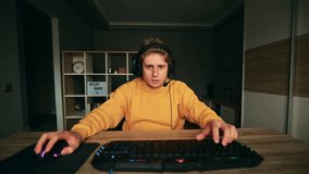 An emotional gamer in a headset plays a video game on a computer at home and loses with a sad face and removes the headphones from his head. The guy lost in an online computer game.