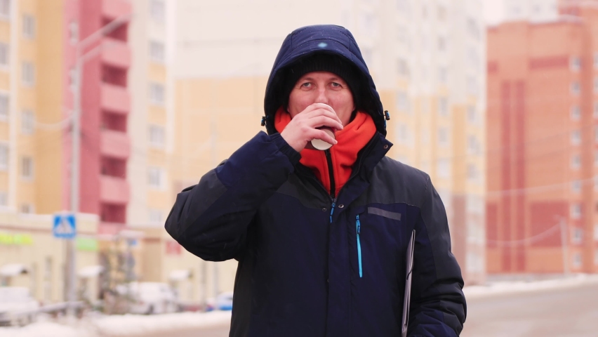 A young man is waiting for a taxi and drinking coffee. Male with a mustache in a warm jacket outdoors in cool weather. In the background, multi-storey high-rise buildings out of focus.