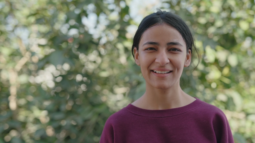 Close static shot of a cheerful and beautiful young Asian Indian female college student with a smile on the face looking at the camera in a broad daylight, green trees in the background Royalty-Free Stock Footage #1068057884