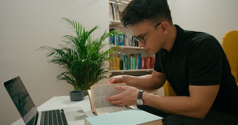Caucasian Christian Pastor Man Studies Holy Bible using Laptop, Book and Notebook at Home. Young Religious Man Participating at Online Conference or Preacher Writing sermon. 4K Medium Zoom out Shot