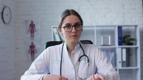 Health concept. Focused Female doctor speaking. Young woman doctor wearing white medical coat, glasses and stethoscope talking on-online video call on laptop while sitting at desk in clinic. Close-up.