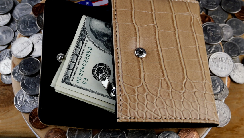 Open wallet from which a few 100 American dollars are sticking out and a lot of scattered American cents are rotate on circle. American flag seen from under purse. Close-up.